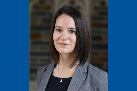 Pictured is Dr. Emma Chory, PhD, a smiling woman with shoulder-length dark brown hair, wearing a gray blazer over a black top and a silver necklace with a small pendant. The background is blurred, featuring an elegant architectural setting with warm tones